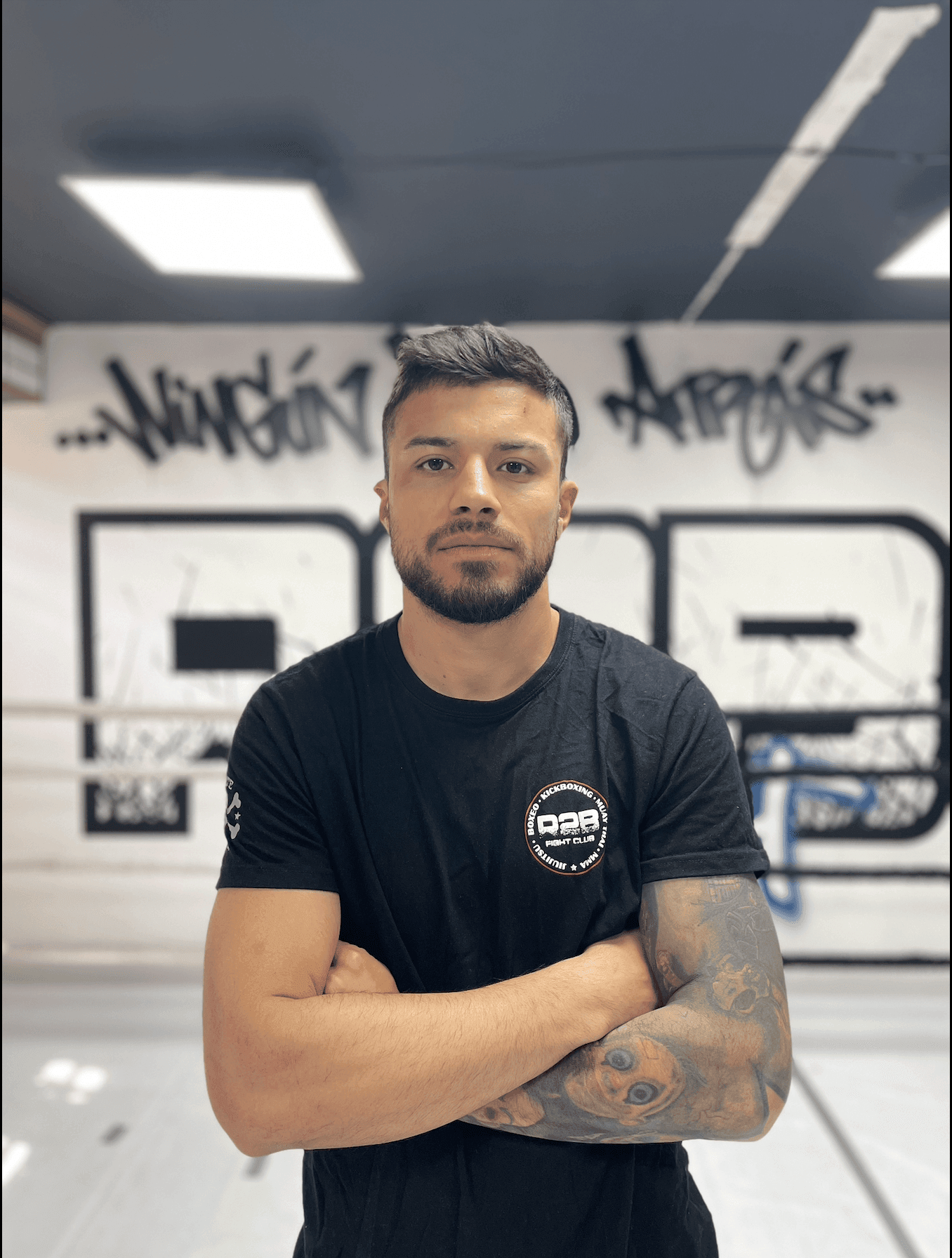 Man with folded arms and tattoos poses in front of a gym wall with graffiti art.